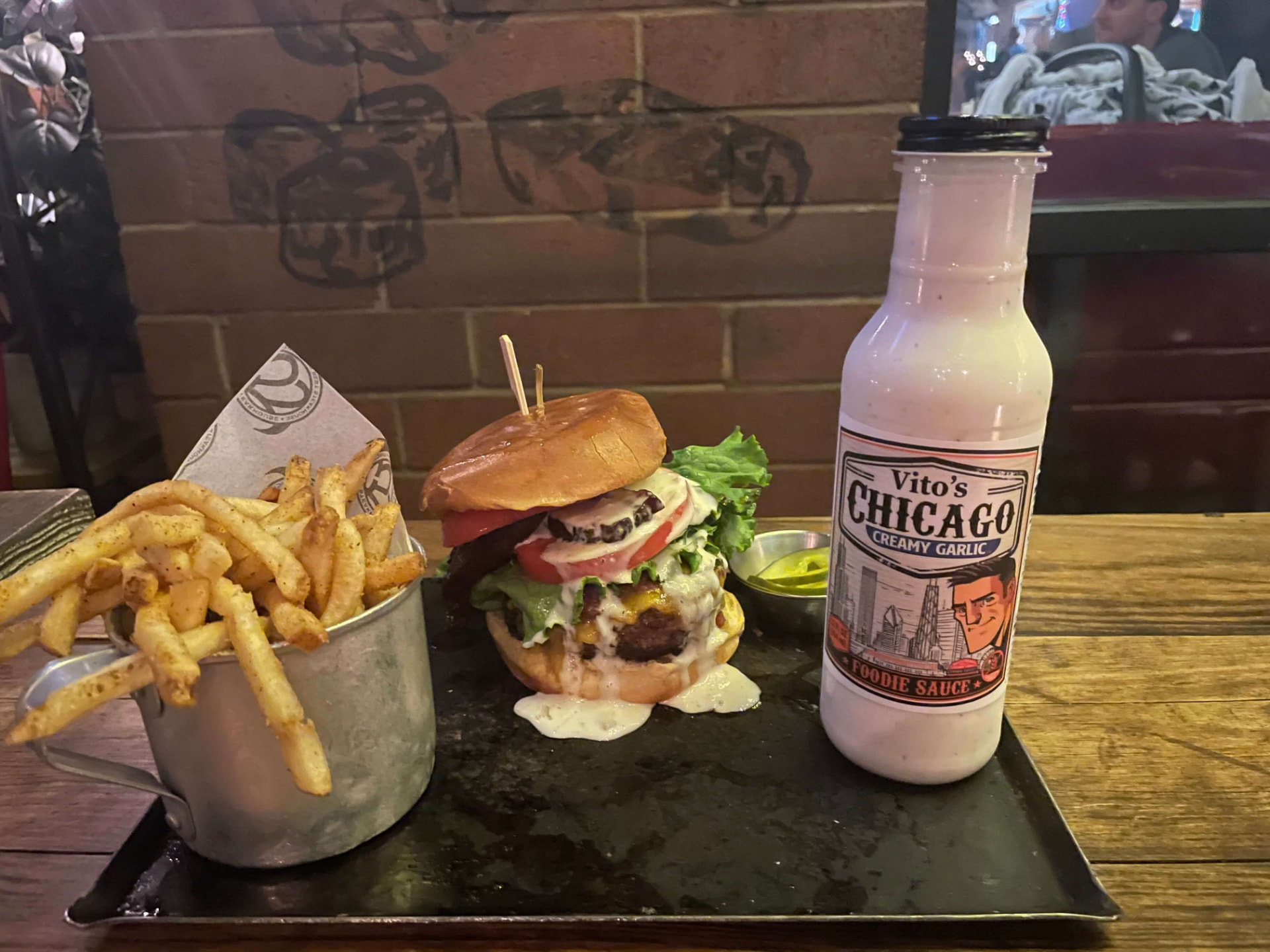 Burger, fries and a bottle of Vito's Chicago Creamy Garlic Foodie Sauce on a slate tray against a brick wall